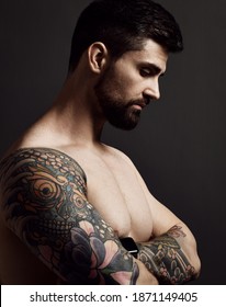 Portrait in profile of naked handsome bearded brutal muscular brunet man with tattoo on arms shoulders holding hands crossed at chest looking down. Hipster stylish look for free lifestyle concept