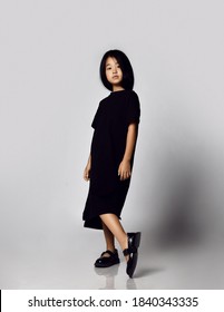 Portrait In Profile Of Self-reliant Asian Kid Girl In Black Long T-shirt Dress And Bob Hairstyle Showing Off Demonstrating Her New Black Sandals Shoes And Looking Down At Camera