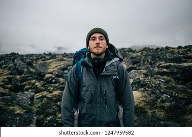 Portrait Of Proud And Brave Young Adventurer Hiker, Explorer Handsome Man With Trekking Backpack Stand On Rocky Hiking Trail Or Path, Look Into Camera. Outdoor Vibes And Adventures Wanderlust