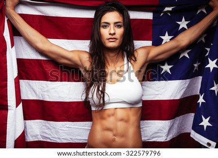 Portrait of proud female athlete holding American Flag against. Muscular young woman looking confidently at camera.
