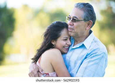 Portrait Proud Hispanic Father Cuddling Happy Relaxed Smiling Attractive Adult Daughter Outdoor, Blurred Background.