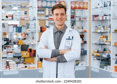 Portrait of a qualified, friendly male pharmacist wearing a white coat, crossing his arms, and looking at the camera, with a shelf of various medicine boxes in background at drugstore or pharmacy. - Powered by Shutterstock