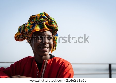 Portrait of a radiant African beauty wearing a red caftan and a colorful headscarf with a traditional pattern sitting casually by the river