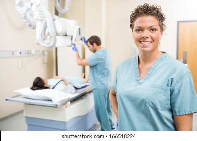 Portrait Of Radiologist Smiling While Colleague Preparing Patient For Xray In Examination Room