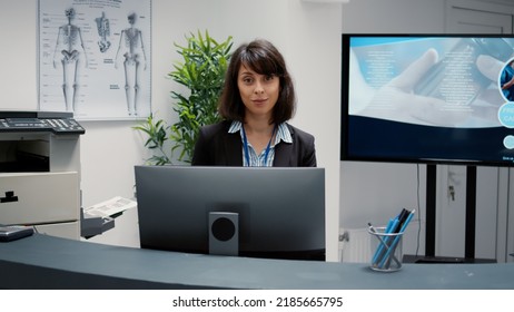 Portrait Of Receptionist Working At Hospital Reception Desk To Help People With Appointment For Checkup Visit. Waiting Area With Administrative Information To Support Healthcare System.