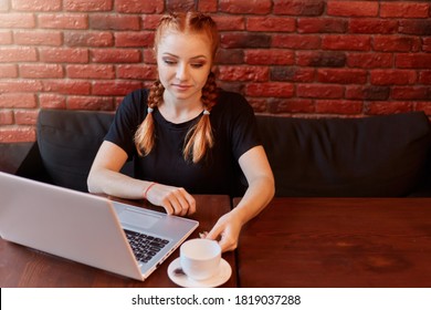 Portrait of red haired coworker business woman working via laptop against brick wall in cafe, concentrated student sitting at table on black sofa, enjoying hot coffee while studying online.