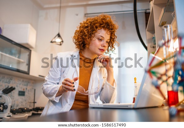 Portrait of red haired female scientist  sitting at desk\
and working with laptop during scientific experiment. Busy\
researcher typing scientific article. Female scientist working on\
laptop 