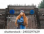 Portrait of a red haired woman in a blue medieval linen dress smiling against the background of a wooden castle gate.