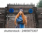 Portrait of a red haired woman in a blue medieval linen dress smiling against the background of a wooden castle gate.