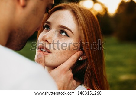 Portrait of a red haired woman with freckles and green eyes looking at her boyfriend while he is touching her lips with a finger against sunset while dating