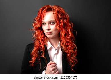 Portrait Of A Red-haired Businesswoman In A White Shirt And Black Jacket Confidently Squeezes Her Fist Looking At The Camera. Confident Young Female Politician On A Dark Background. Election Promise.