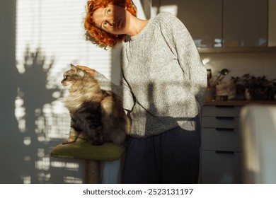 portrait of red-haired curly young woman, in home kitchen, with pet gray fluffy cat in her arms, in sun rays and shadow, lit by bright sun - Powered by Shutterstock