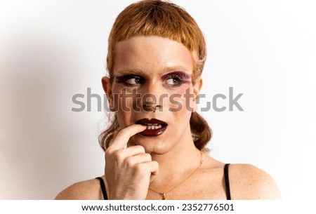 A portrait of a red-haired non-binary person looking to the side while biting a nail on a white background. Concept of non-binary people, nervous, restless. Person does not identify with any gender.