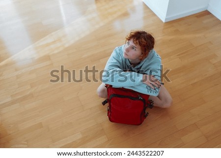 portrait red-haired teenage girl sitting on parquet floor, with suitcase, along in an empty apartment. concept moving, rent, student life