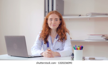 Portrait Redhead Doctor Sitting At The Desk In Office. Candid Professional Woman Therapist Posing Looking At The Camera And Friendly Smiling.