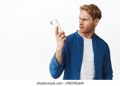 Portrait Of Redhead Guy Looks Confused At Smartphone, Hear Something Strange In Mobile Phone During Call, Standing Puzzled Against White Background