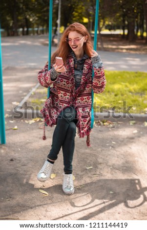 Portrait of redhead young beautiful woman with freckles and dark lipstick wearing sunglasses sitting on swing on playground outdoors in park using mobile phone.