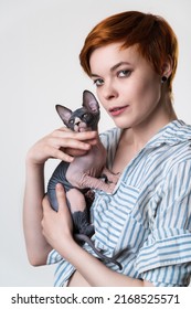 Portrait Of Redhead Young Woman Holding Sphynx Cat In Hands And Looking At Camera. Hipster Female With Short Hair Dressed In Striped White-blue Shirt. Studio Shot On White Background. Part Of Series.