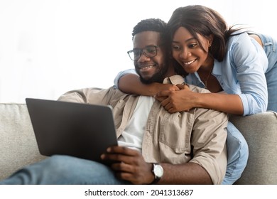 Portrait Of Relaxed African American Young Family Using Digital Tablet Resting Together In Living Room, Woman Hugging Her Husband Sitting On Couch. Happy Black Couple Looking At Pad Screen