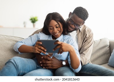 Portrait Of Relaxed African American Young Family Using Digital Tablet Together Resting On Couch In Living Room, Male In Glasses And Braces Hugging Female. Happy Black Couple Looking At Pad Screen