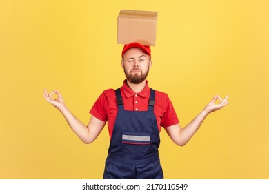 Portrait Of Relaxed Courier Man Standing With Cardboard Box On His Head, Practicing Yoga, Looking At Camera With One Eye, Calms Down. Indoor Studio Shot Isolated On Yellow Background.