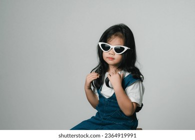 Portrait of relaxed cute little asian girl child funny smiling and laughing wearing white headphones and glasses, keeps hands on her ears, posing and smiling enjoying isolated over gray background. - Shutterstock ID 2364919991