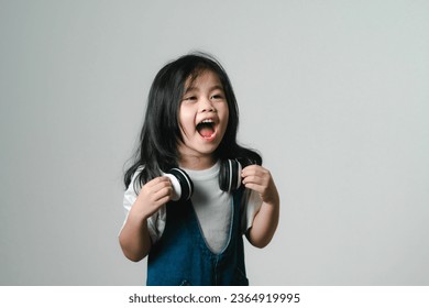 Portrait of relaxed cute little asian girl child funny smiling and laughing wearing white headphones, keeps hands on her ears, posing and smiling enjoying isolated over gray background. - Shutterstock ID 2364919995