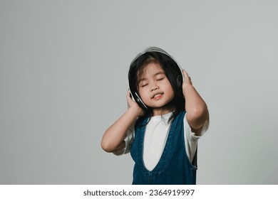 Portrait of relaxed cute little asian girl child wearing white headphones, keeps hands on her ears, posing and smiling enjoying isolated over gray background. - Shutterstock ID 2364919997