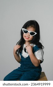 Portrait of relaxed cute little asian girl child funny smiling and laughing wearing white headphones and glasses, keeps hands on her ears, posing and smiling enjoying isolated over gray background. - Shutterstock ID 2364919999