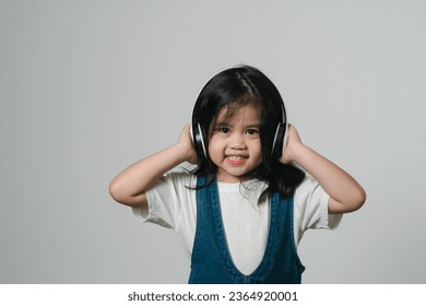 Portrait of relaxed cute little asian girl child funny smiling and laughing wearing white headphones, keeps hands on her ears, posing and smiling enjoying isolated over gray background. - Shutterstock ID 2364920001