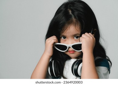 Portrait of relaxed cute little asian girl child funny smiling and laughing wearing white headphones and glasses, keeps hands on her ears, posing and smiling enjoying isolated over gray background. - Shutterstock ID 2364920003