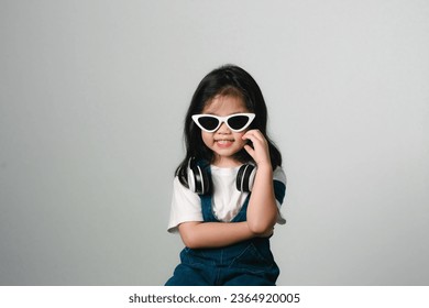 Portrait of relaxed cute little asian girl child funny smiling and laughing wearing white headphones and glasses, keeps hands on her ears, posing and smiling enjoying isolated over gray background. - Shutterstock ID 2364920005