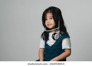 Portrait of relaxed cute little asian girl child funny smiling and laughing wearing white headphones and glasses, keeps hands on her ears, posing and smiling enjoying isolated over gray background. - Shutterstock ID 2364920015