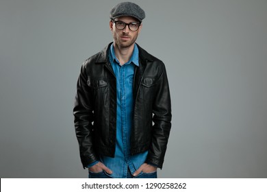 portrait of relaxed fashion man wearing flat cap and leather jacket standing with hands in pockets on light grey background