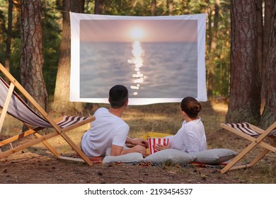 Portrait Of Relaxed Loving Couple Lying In The Forest With Overhead Projector, Watching Romantic Movie, Enjoying Sunset On Screen And Beautiful Nature In The Wood.