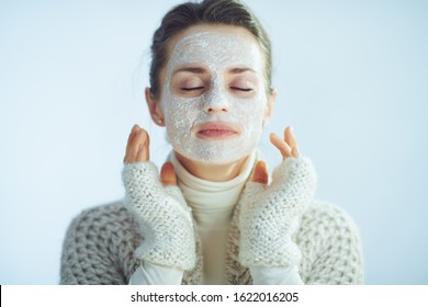 Portrait Of Relaxed Modern Woman In Roll Neck Sweater And Cardigan With White Facial Mask As Part Of Winter Skin Care Against Winter Light Blue Background.