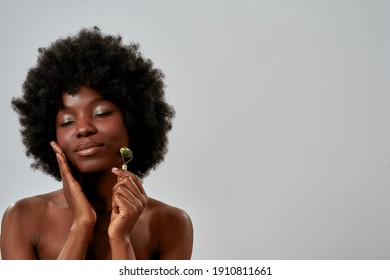 Portrait of relaxed young african american woman with perfect glowing skin massaging her face with jade roller, posing with eyes closed isolated over gray background. Beauty, skin care concept