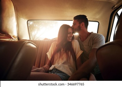 Portrait of relaxed young couple on a road trip sitting in back seat of car. Man kissing forehead of his girlfriend, with bright sunshine from behind.
