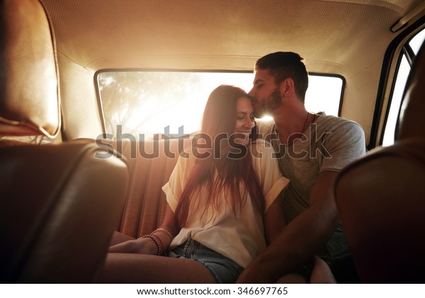 Portrait of relaxed young couple on a road trip\
sitting in back seat of car. Man kissing forehead of his\
girlfriend, with bright sunshine from\
behind.