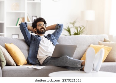 Portrait Of Relaxed Young Indian Man With Laptop Sitting On Couch At Home Interior, Smiling Eastern Freelancer Guy Leaning Back On Couch With Hands Behind Head, Having Break In Online Work - Powered by Shutterstock