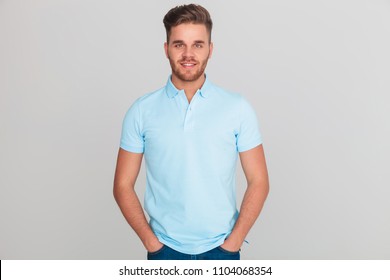 Portrait Of Relaxed Young Man Wearing Light Blue Polo T-shirt While Standing On Light Grey Background With Hands In Pockets