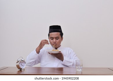 Portrait Of Religious Asian Man In Koko Shirt Or White Muslim Shirt And Black Cap Eating Instant Noodles For Suhoor To Be Strong During Fasting From Dawn Until Dusk In Ramadan Month