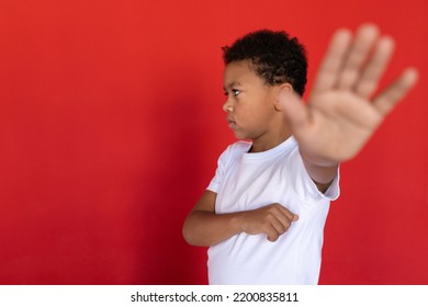 Portrait Of Resentful Preteen Boy Making Stop Gesture. Mixed Race Child Wearing White T-shirt Looking Away And Stretching Arm Against Red Background. Abuse And Home Violence Concept