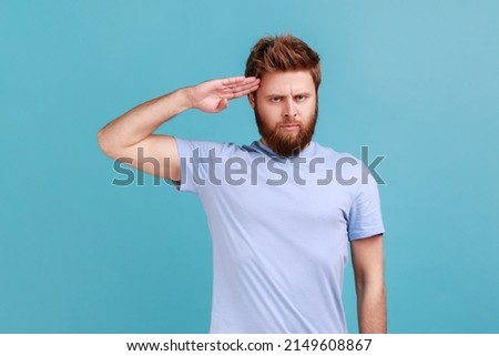 Portrait of responsible patriotic bearded man saluting with respect as if soldier waiting order from commander, obeying discipline. Indoor studio shot isolated on blue background.