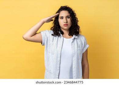 Portrait of responsible serious woman with dark wavy hair saluting commander, listening order with obedient expression, looking at camera. Indoor studio shot isolated on yellow background.