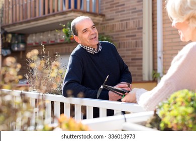 Portrait Of Retired Couple On A Terrace: Old Husband Is Holding A Cup Of Hot Beverage And Talking To His Wife Who Is Holding Gardening Tools