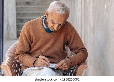 Portrait of retired Indian senior man writing in diary while sitting on chair at home. Old people hobby concept. 
