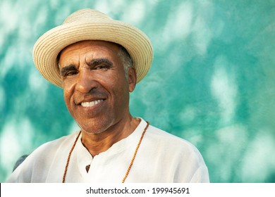 Portrait Of Retired Senior Hispanic Man With Straw Hat Sitting In Park And Looking At Camera With Happy Expression