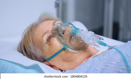 Portrait Of Retired Senior Woman Breathing Slowly With Oxygen Mask During Coronavirus Covid-19 Outbreak. Old Sick Lady Lying In Hospital Bed, Getting Treatment For Deadly Infection