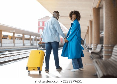 Portrait Of Romantic Black Couple Walking With Suitcase At Railway Station, Happy African American Spouses Holding Hands And Turning At Camera While Going To Train Departure, Copy Space